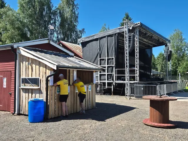 Två funktionärer vid jetongkiosken med scenen i bakgrunden en solig dag.