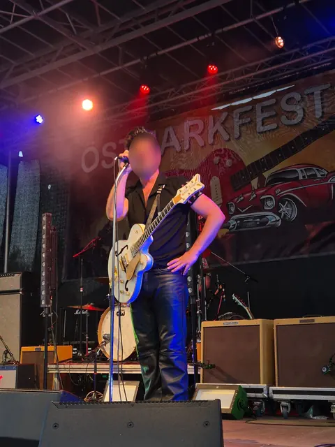 Sångare med vit Gretsch-gitarr framför festivalens banderoll med röd gitarr och amerikansk veteranbil.
